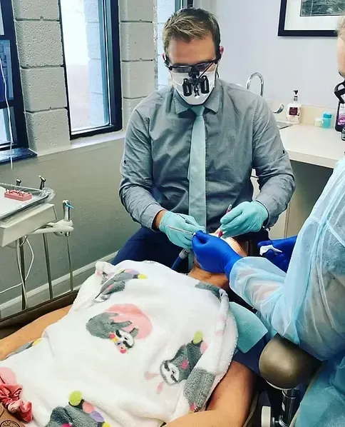 Dentist wearing loupes and mask examines a patient covered with a sloth-print blanket.